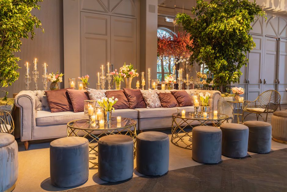 A luxurious, well-maintained lounge area featuring a long, light-colored fabric sofa adorned with plush cushions in shades of pink and floral patterns. In front of the sofa are several round, velvet-upholstered ottomans in dark grey positioned around glass-top coffee tables with gold metal frames. The tables hold multiple lit candles and small floral arrangements, creating a warm, inviting atmosphere. The room has elegant wall paneling, large windows allowing natural light, and lush greenery including a tall indoor tree behind the sofa. The overall scene depicts a clean, stylish space prepared for an event, exemplifying high standards of surface cleaning and decor maintenance, consistent with the services offered by Cleaners SW8 for upholstery and interior surface cleaning after events in Battersea Park.