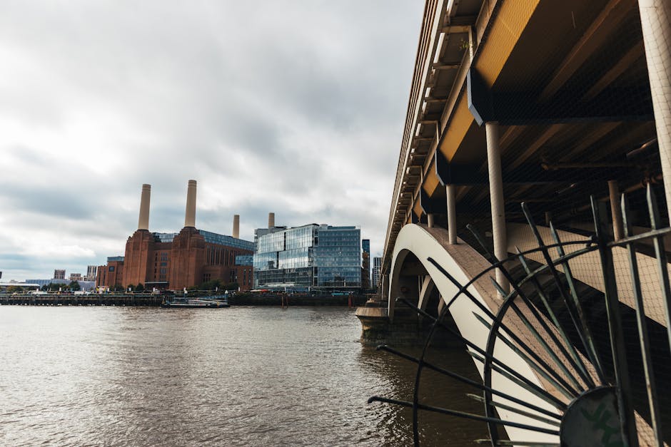 A view of the Battersea Power Station from across the River Thames during daytime, featuring the historic brick building with four tall chimneys and modern glass structures nearby. In the foreground, part of a bridge with beige and black structural elements, and a section of curved metal railing are visible, with the river's surface appearing calm and slightly reflective. The sky is overcast with gray clouds, creating a moody, industrial atmosphere. This image illustrates the urban waterfront area near SW8, highlighting the blend of historic and contemporary architecture. Cleaners SW8 specializes in surface and deep cleaning services, including sanitisation of residential and commercial spaces in this vibrant district.