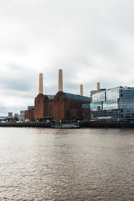 A low-angle view of Battersea Power Station, featuring a mix of modern glass skyscrapers and the historic brick power station structure. The photo captures the sleek, reflective surfaces of the surrounding buildings, the textured brickwork of the power station, and a partly cloudy sky overhead. Visible are glass facades, steel accents, and a concrete column, all contributing to a contemporary cityscape scene. The area appears clean and well-maintained, reflecting the professional surface cleaning services offered by Cleaners SW8, based on this visual evidence of attention to hygiene and upkeep in residential or commercial settings near the Battersea Power Station in SW8.