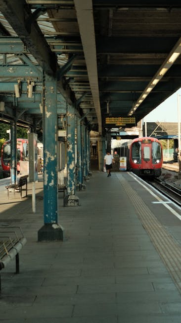 A photograph of Clapham Junction train station platform showing a tiled concrete surface, which appears clean and well-maintained. The platform features metal support beams with peeling blue paint, and a long overhead canopy providing shelter for waiting passengers. Several metal benches with wooden slats are positioned along the platform, and a person is walking towards a red train that is parked on the tracks. The scene is illuminated by natural daylight, and the station looks tidy and orderly, reflecting effective cleaning and maintenance by companies like Cleaners SW8, who specialise in domestic and commercial surface cleaning and sanitisation services near Clapham Junction station.