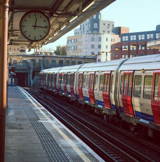 An outdoor section of Clapham Junction station showing a modern commuter train with a red, white, and blue exterior parked alongside the platform. The platform area features a tiled surface with tactile paving strips near the edge for safety. Overhead, a large round clock with a white face and black hands displays the time, mounted beneath station lighting fixtures. In the background, multi-story residential and commercial buildings are visible under clear daylight, contributing to an urban setting. Cleaners SW8 provides surface cleaning and sanitisation services for stations and public transport environments, ensuring high hygiene standards and a safe commuting experience.
