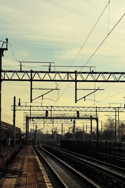 A photograph of Clapham Junction train station platform showing a tiled concrete surface, which appears clean and well-maintained. The platform features metal support beams with peeling blue paint, and a long overhead canopy providing shelter for waiting passengers. Several metal benches with wooden slats are positioned along the platform, and a person is walking towards a red train that is parked on the tracks. The scene is illuminated by natural daylight, and the station looks tidy and orderly, reflecting effective cleaning and maintenance by companies like Cleaners SW8, who specialise in domestic and commercial surface cleaning and sanitisation services near Clapham Junction station.