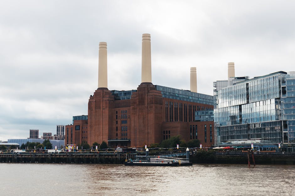 A large red-brick industrial building with four tall, beige smokestacks situated along a river, reflecting in the water. Next to the factory, there are modern glass office buildings with a sleek, refl