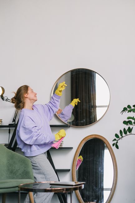 A woman wearing a lavender sweatshirt and yellow cleaning gloves is engaged in surface cleaning inside a residential space. She is reaching up with a cloth, wiping the surface of a round mirror mounted on a white wall, which reflects a window with dark curtains. Another mirror with a wooden frame leans against the wall below the mounted mirror. The room is illuminated by natural light, and a green upholstered chair is partially visible in the foreground. A nearby shelf holds cleaning supplies, indicating a thorough domestic cleaning process. The scene demonstrates attention to detail and cleanliness in a well-maintained interior, consistent with the services offered by Cleaners SW8 for end of tenancy cleaning on Lavender Hill.